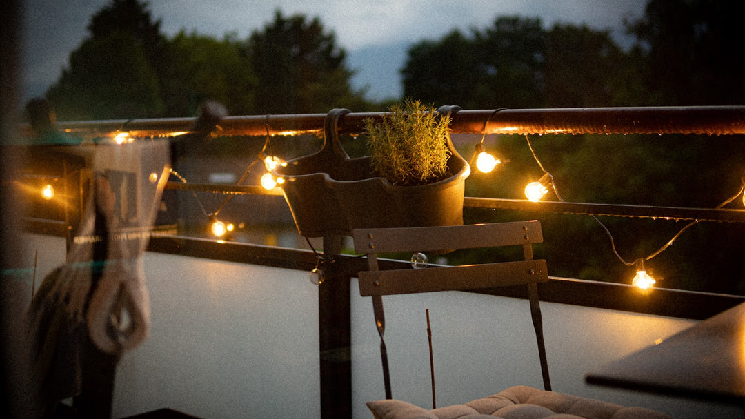 Balkon in der Dämmerung mit warmer Lichterkette am Geländer, Kräutertopf und Stuhl mit Sitzkissen