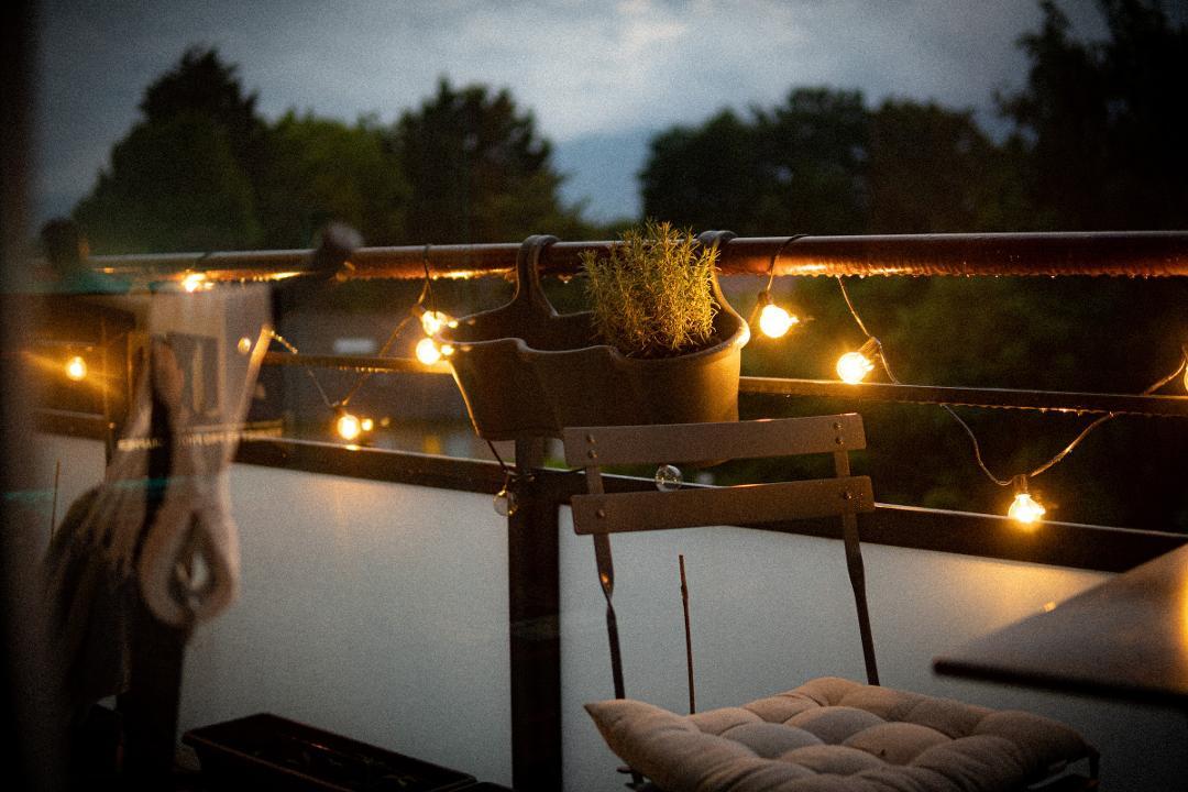 Balkon in der Dämmerung mit warmer Lichterkette am Geländer, Kräutertopf und Stuhl mit Sitzkissen