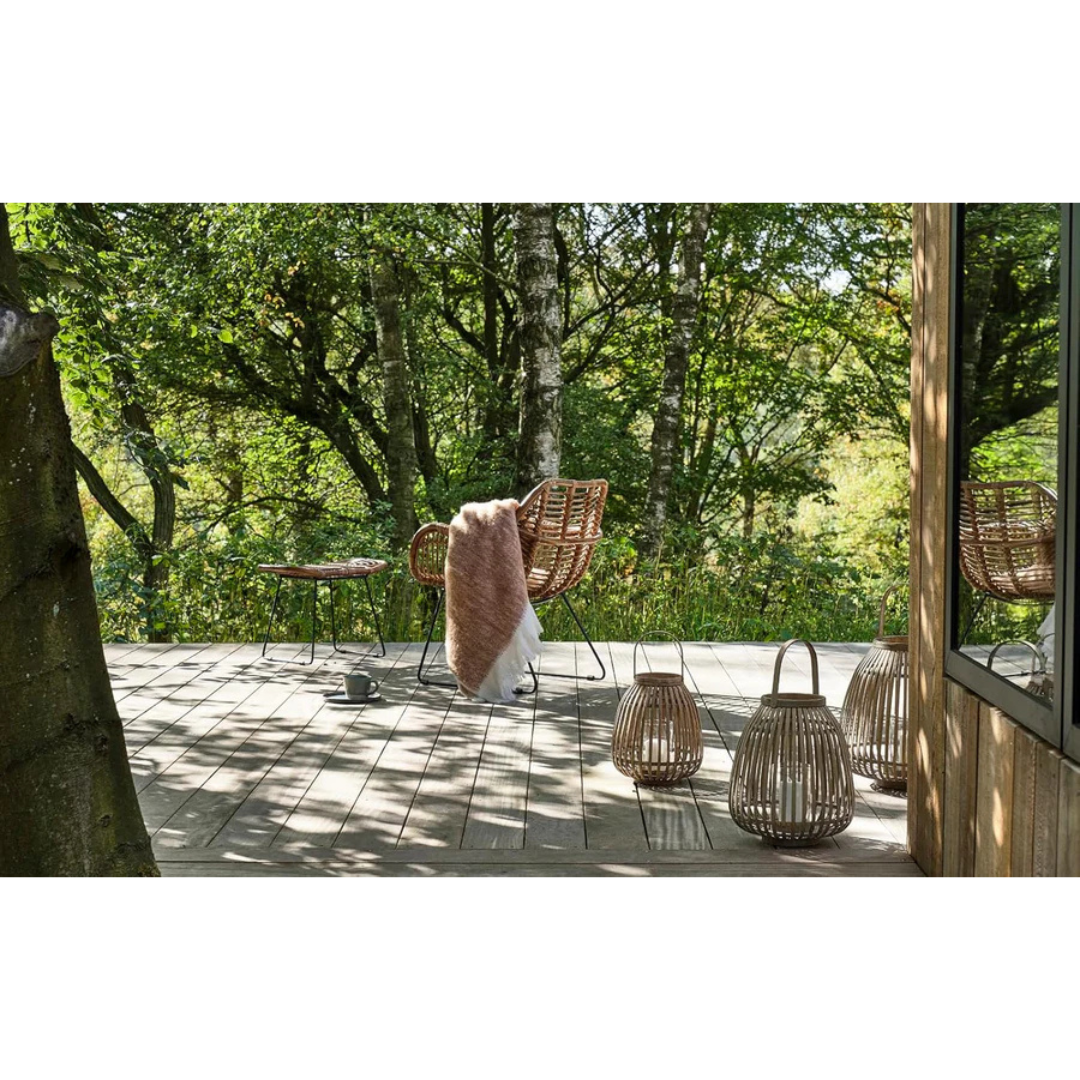 Bambus-Laternen auf einer Holzterrasse vor Bäumen, daneben Rattansessel mit Decke