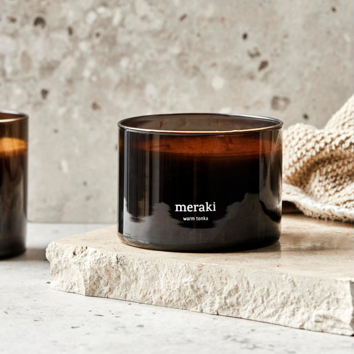 Bougie parfumée Meraki Warm Tonka, sur une dalle en pierre à côté d'un plaid tricoté